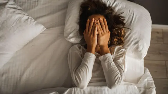 Uma mulher cobrindo o rosto com as mãos deitada na cama